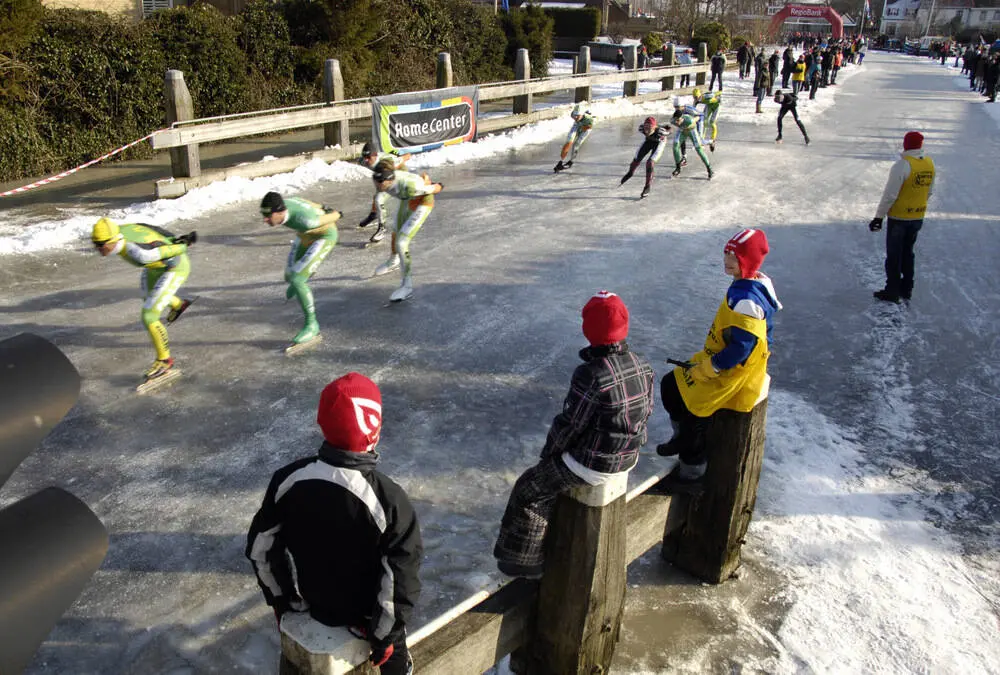 Drie jongetjes volgen de schaatsmarathon van Akkrum op de voet, vanaf de dukdalven van een van de brugen in het dorp, die speciaal voor de schaatswedstrijd ruim een uur open stonden. Winnaar werd Jorrit Bergsma uit het even verderop gelegen Aldeboarn. Op het bevroren riviertje de Boarne schaatste hij een thuiswedstrijd.